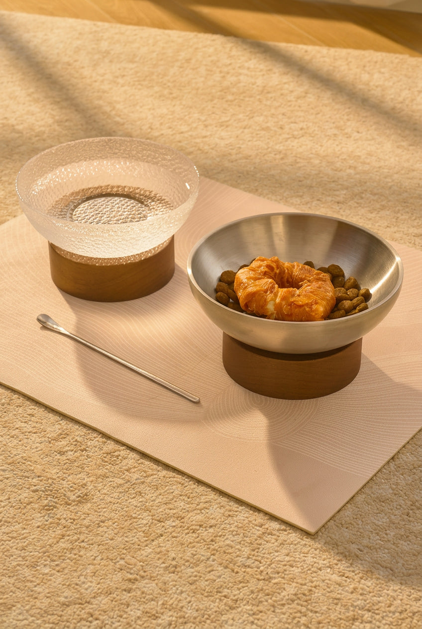 A pet elevated food bowl set on a beige textured rug, featuring a stainless steel bowl with a wooden base containing wet and dry food, and a matching clear glass water bowl on a wooden base. A metal spoon rests beside the bowls.